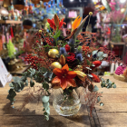 Prachtig kleurrijk boeket met rode en oranje bloemen, blauwe distels en gouden accenten, gepresenteerd in een glazen vaas op een houten tafel.