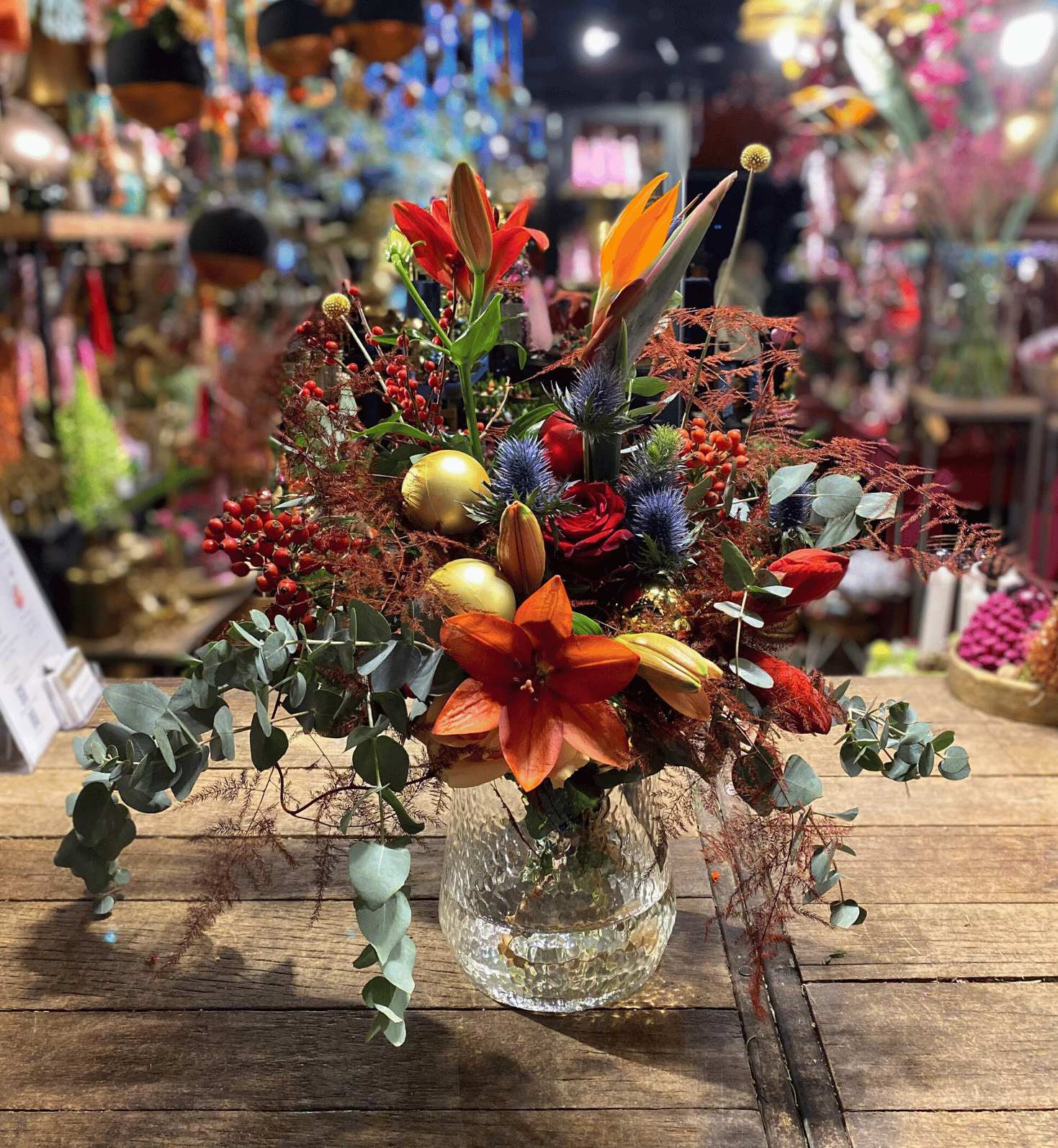 Prachtig kleurrijk boeket met rode en oranje bloemen, blauwe distels en gouden accenten, gepresenteerd in een glazen vaas op een houten tafel.